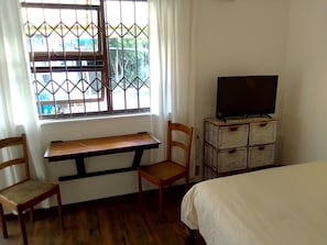 Quarto com escrivaninha de madeira, duas cadeiras de madeira, televisão sobre um suporte e uma cama.