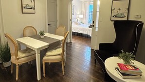 A room with a dining table, chairs, a black armchair, a small table with a book and plant, and a bed in the background. - Exc Location & Comfortable Apt 2BD 1BA Downtown JC 12 min to NYC (Jersey City)