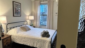 A bedroom with a bed, a bedside table with a lamp, a window with curtains, and a framed picture on the wall. - Exc Location & Comfortable Apt 2BD 1BA Downtown JC 12 min to NYC (Jersey City)