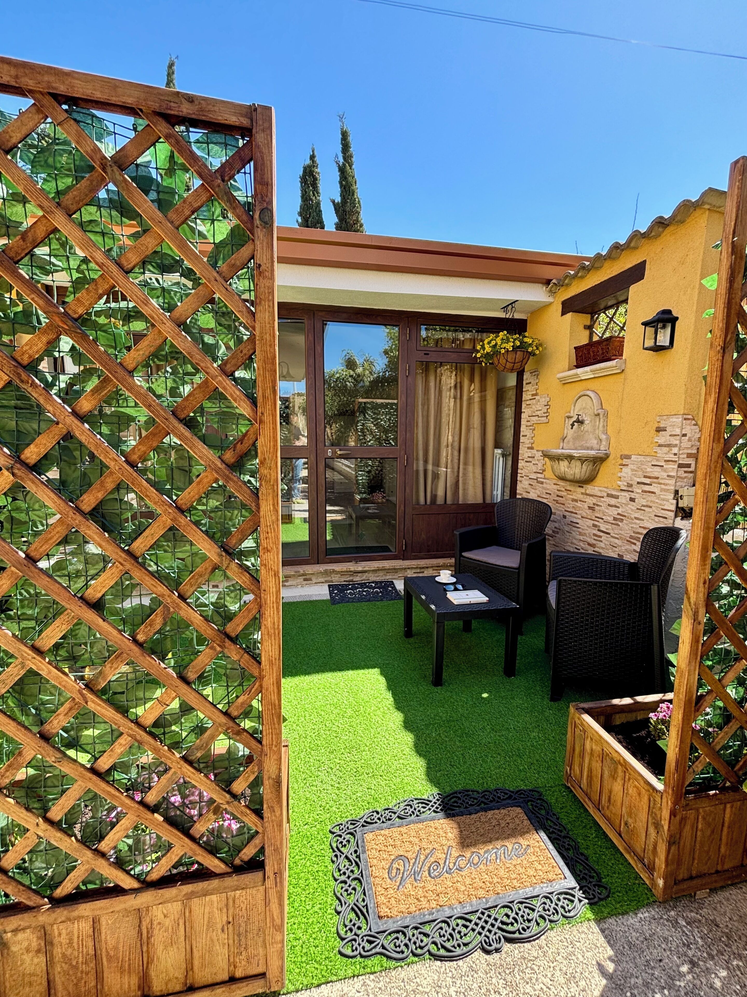 Un espace patio agrémenté d’une treille en bois, d’un paillasson, d’une table avec des chaises et d’un mur en pierre doté d’une fenêtre et d’une lanterne.