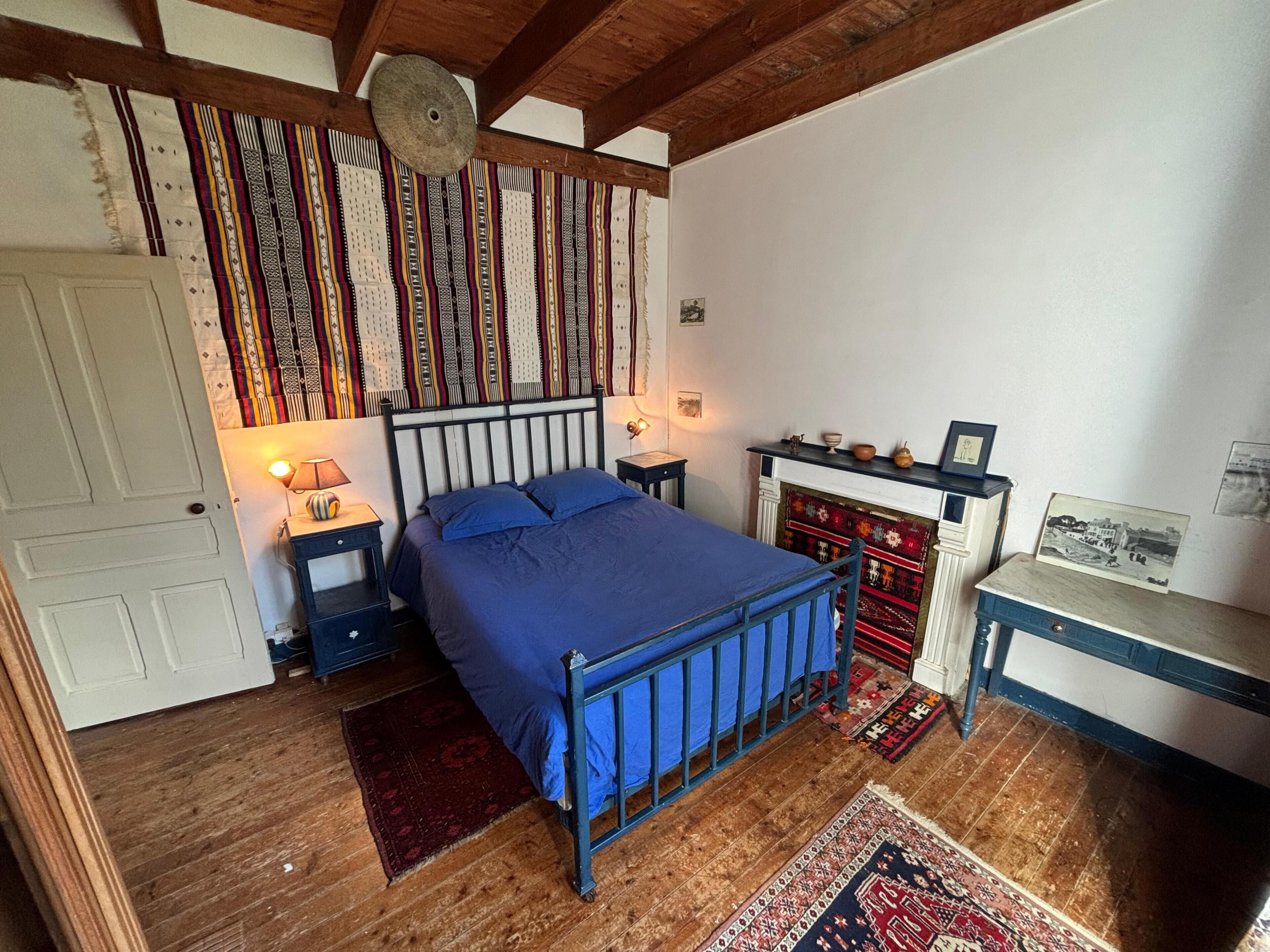 A bedroom with a bed, wooden floor, a fireplace, a nightstand, and a wall with patterned textiles.