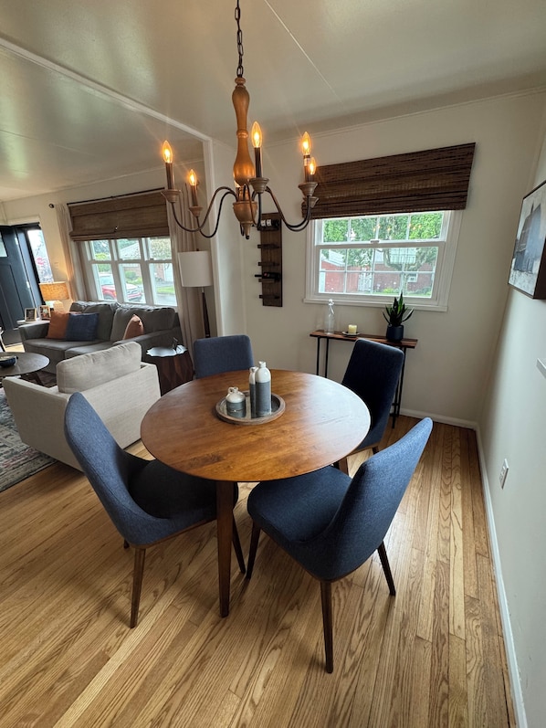 A dining area with a round wooden table, blue upholstered chairs, a chandelier, and a window with a view of the outside.