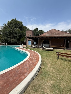 A swimming pool with a brick edge, a covered patio area with outdoor furniture, and a building with a tiled roof.