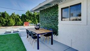A patio with a wooden table and chairs, a hedge wall, and a window. - Vista Ashby: Stagecoach Oasis, Pool & Mtn Views (Palm Springs)