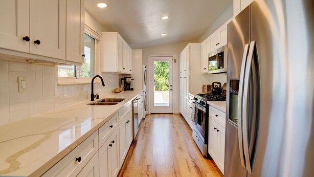 A kitchen with white cabinets, stainless steel appliances, a wooden floor, and a view through a door. - Vista Ashby: Stagecoach Oasis, Pool & Mtn Views (Palm Springs)