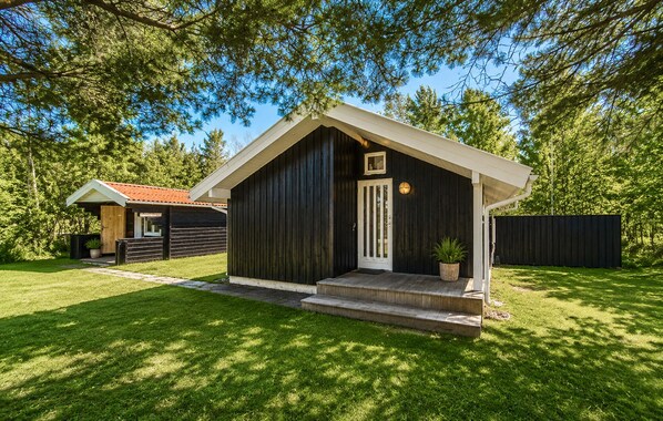 En liten, mörk trästuga med en vit dörr och ett litet fönster, omgiven av en välskött gräsmatta och träd.