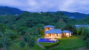 Une maison à deux étages avec une piscine, entourée d’une végétation luxuriante et de collines.