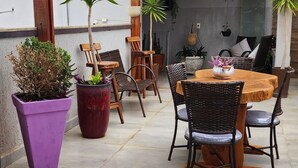 Un patio exterior con una pérgola de madera, una mesa redonda con sillas, plantas en macetas y una zona de estar con cojines.