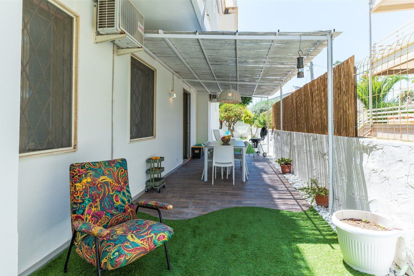 Une terrasse avec une table et des chaises, une chaise aux couleurs vives, un barbecue et un petit coin jardin.