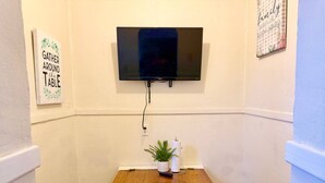 A room with a TV mounted on the wall, a wooden table with a small plant, and a sign that says 'Gather Around the Table'. - Large Group Stay Near Syracuse University & Dome (Syracuse)