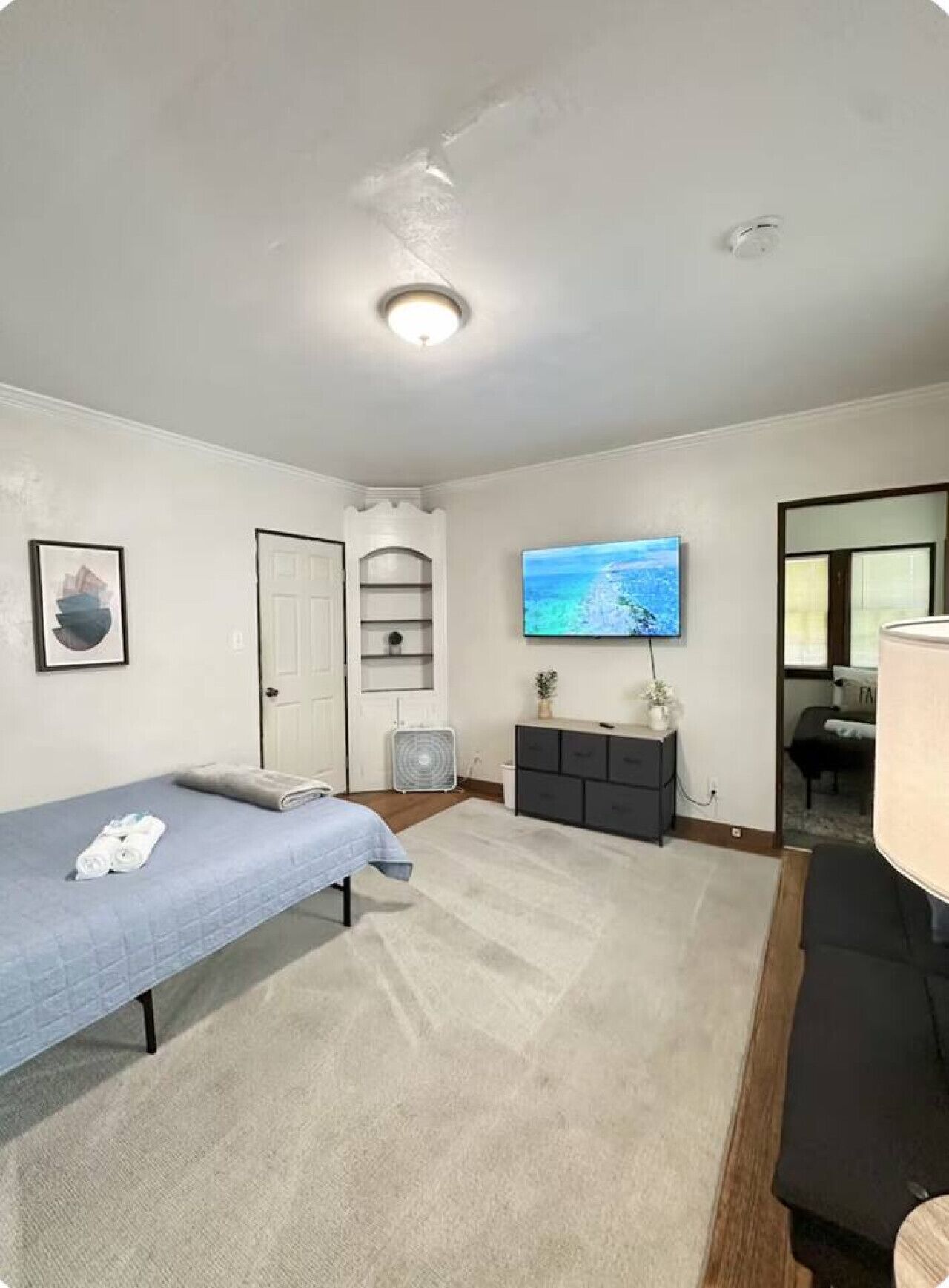 Un dormitorio con una cama individual, televisión, escritorio pequeño, ventilador y puerta que da a otra habitación.