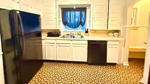 A compact kitchen with white cabinets, a black refrigerator, a dishwasher, and a window with a view of trees. - Large Group Stay Near Syracuse University & Dome (Syracuse)