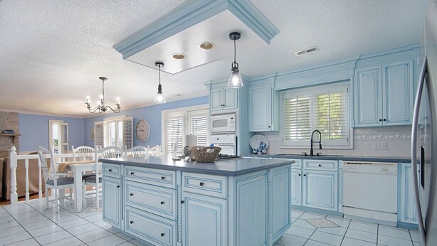 A kitchen with light blue cabinets, a central island, a dining area with white chairs, and a chandelier.