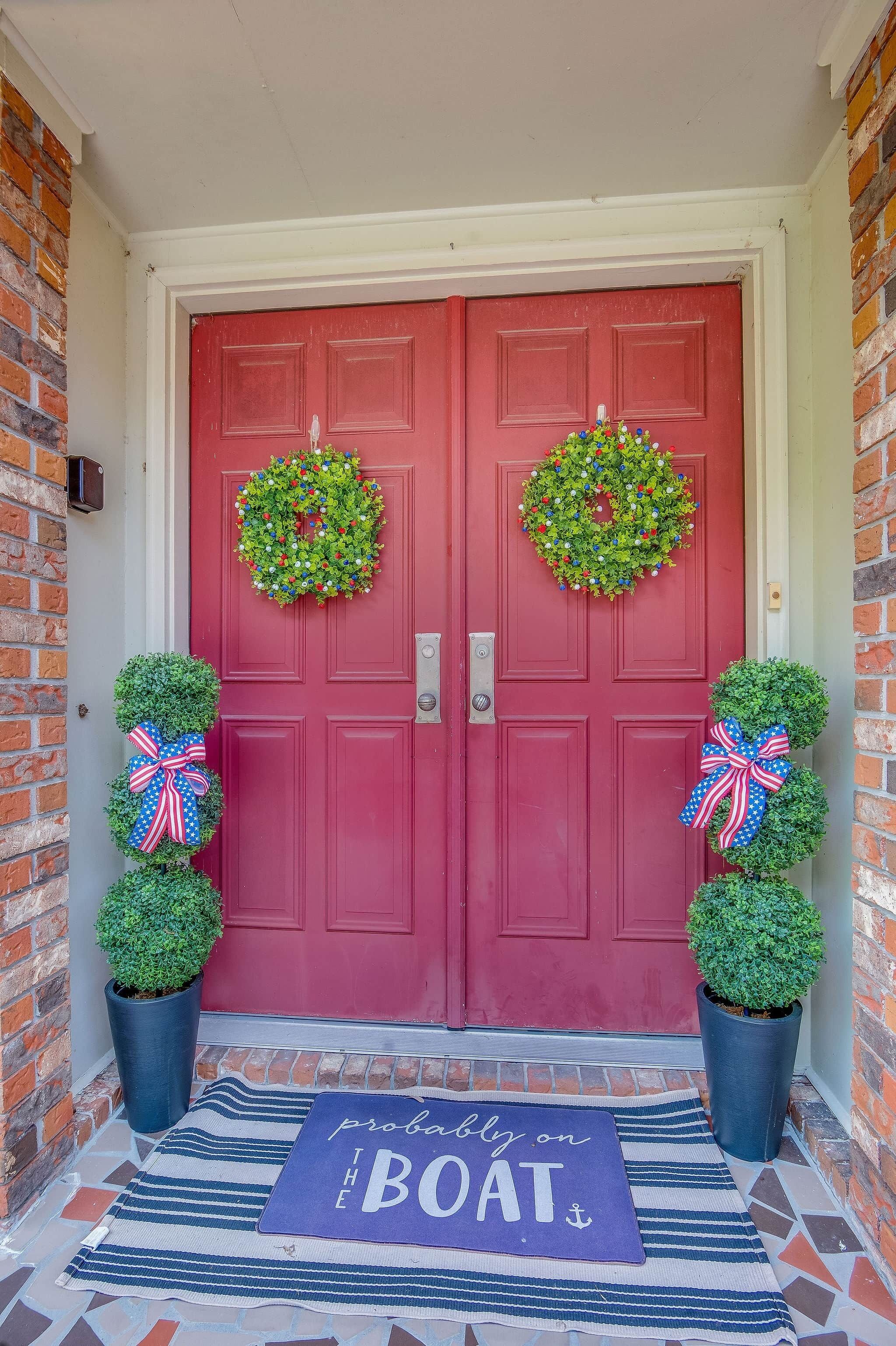 A red door with two wreaths, two potted plants with bows, and a mat with text.