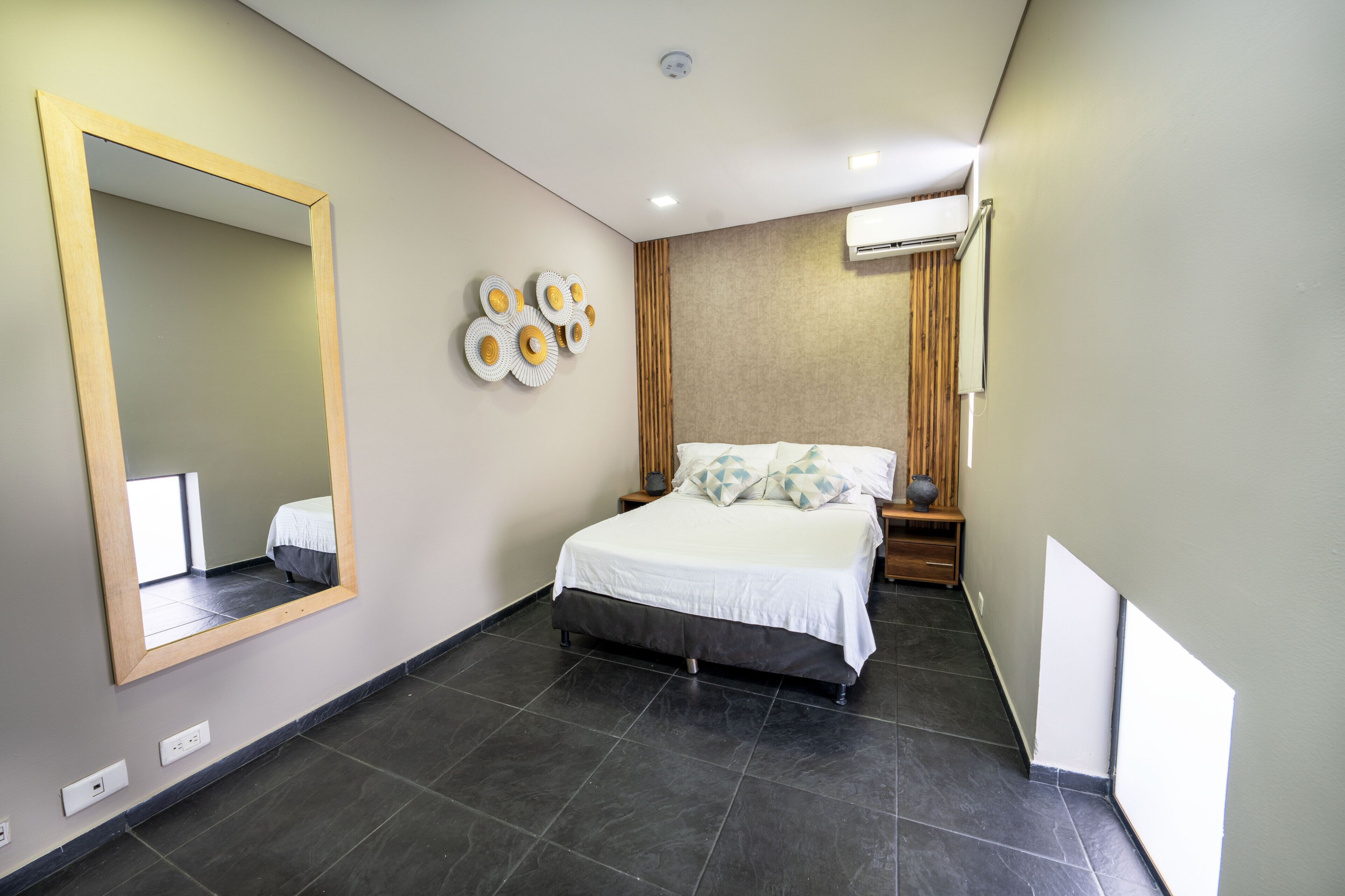 A modern hotel room with a large mirror, a bed with white bedding, a wall-mounted air conditioner, and a decorative wall piece.