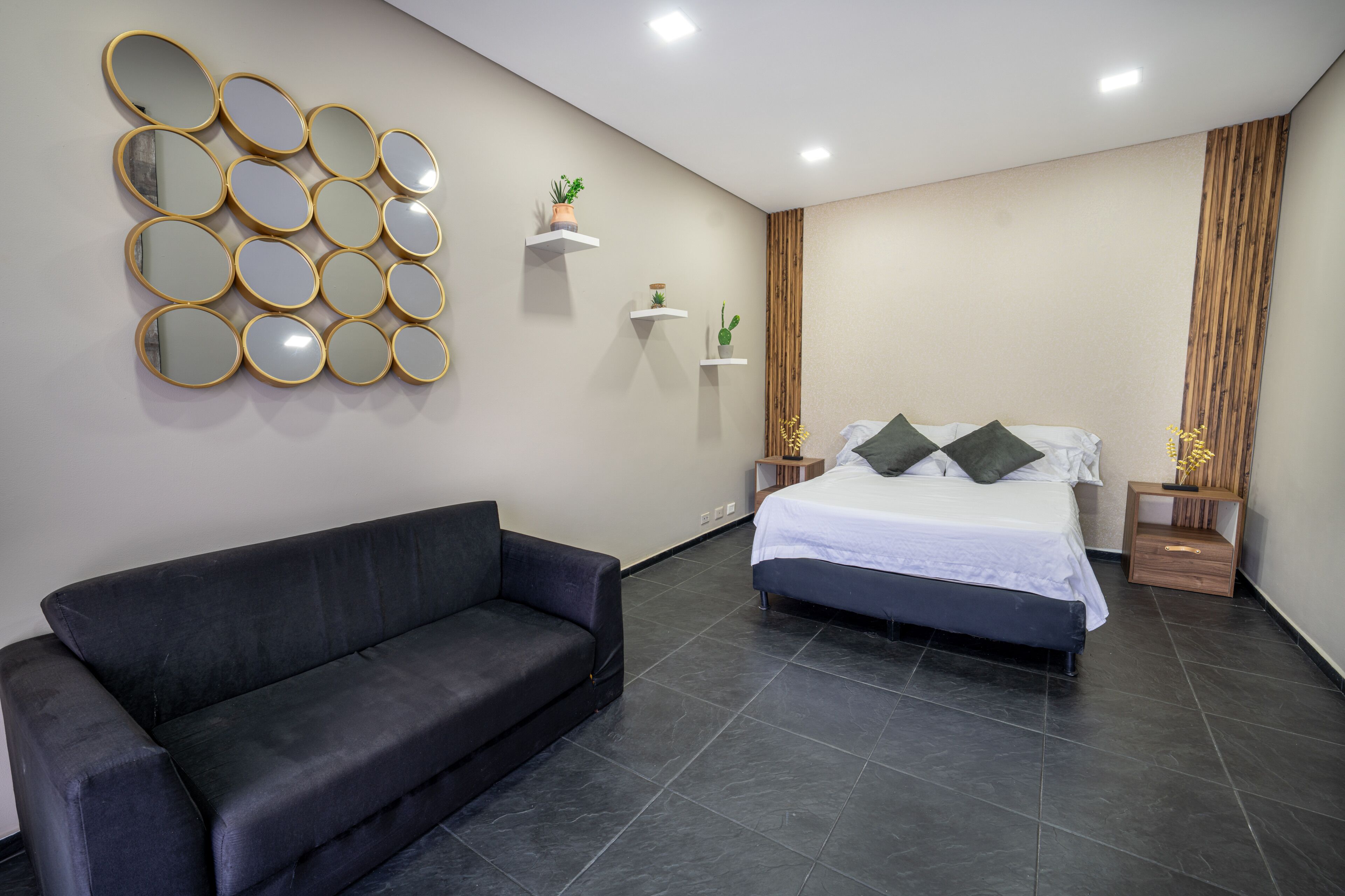Een moderne slaapkamer met een zwarte bank, een bed met witte beddengoed, meerdere spiegels aan de muur en kleine plankjes met planten.