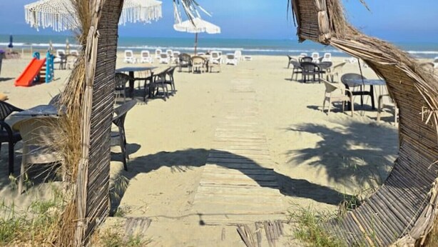 Ein Strand mit einer Reetbogenkonstruktion, Tischen, Stühlen und einem strahlend blauen Himmel.