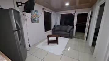 A living room with a flat-screen TV mounted on the wall, a small wooden table, a sofa, and a refrigerator.