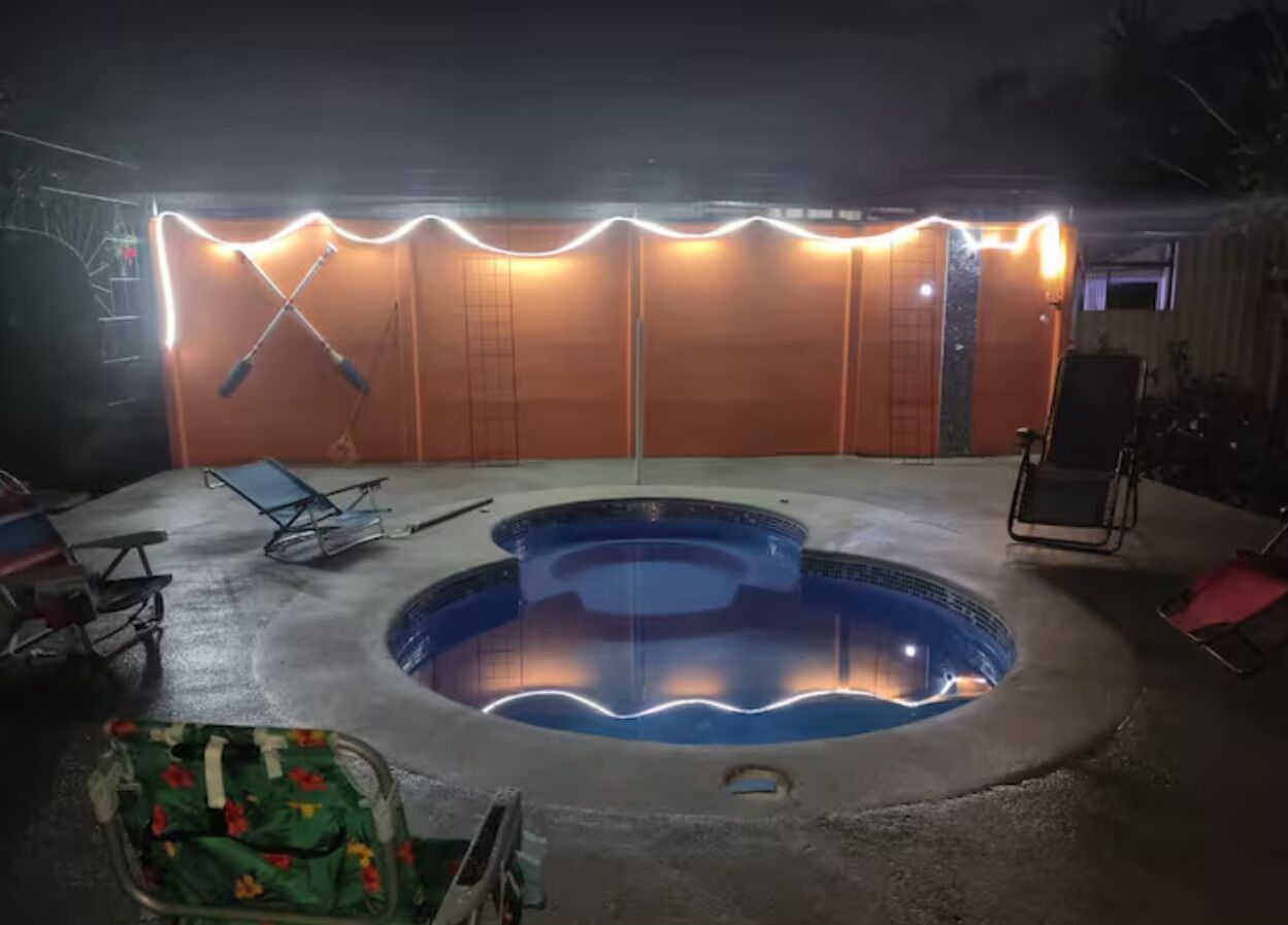A swimming pool with a hot tub, surrounded by lounge chairs, and a covered area with lights.