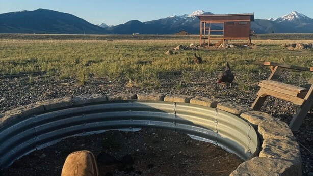 A person relaxing by a fire pit with a view of mountains and a wooden structure in the distance.