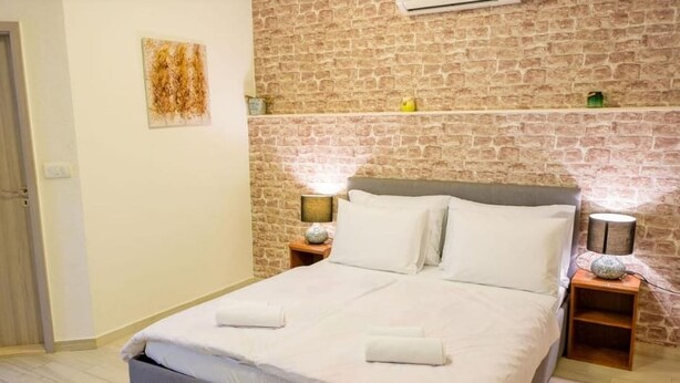 Un lit double avec du linge de lit blanc, deux tables de chevet avec des lampes, un mur décoré d’un motif en briques et une petite étagère avec des objets décoratifs.
