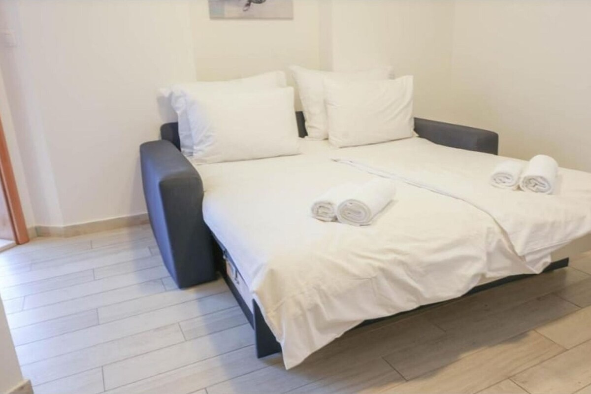 Ein ordentlich bezogenes Bett mit weißen Leinen und Handtüchern, ein dunkelgraues Sofa und ein heller Holzboden.