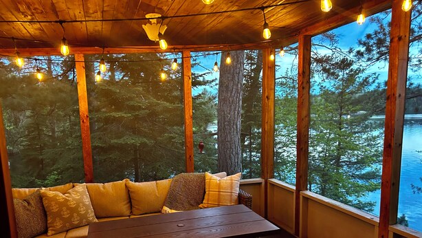 Una terraza de madera con un banco y una mesa, ventanas amplias con vistas al bosque y luces colgantes.