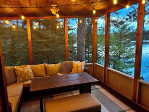 Una terraza de madera con un banco y una mesa, ventanas amplias con vistas al bosque y luces colgantes.