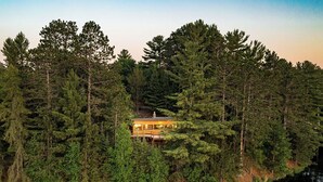 Una cabaña rodeada de un bosque frondoso, con una terraza visible y una chimenea.