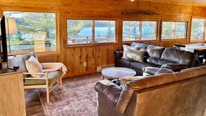 Interior de una cabaña con paredes de madera, un sofá de cuero, una mesa de comedor y ventanas grandes con vistas a un lago.