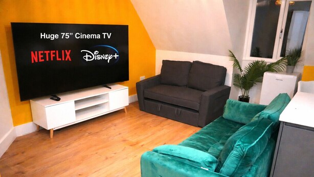 A living room with a large flat-screen TV, a sofa, a small cabinet, and a potted plant. - Super Central 2 Bedroom Apartment Sleeps 5, 85` TV (Canterbury)