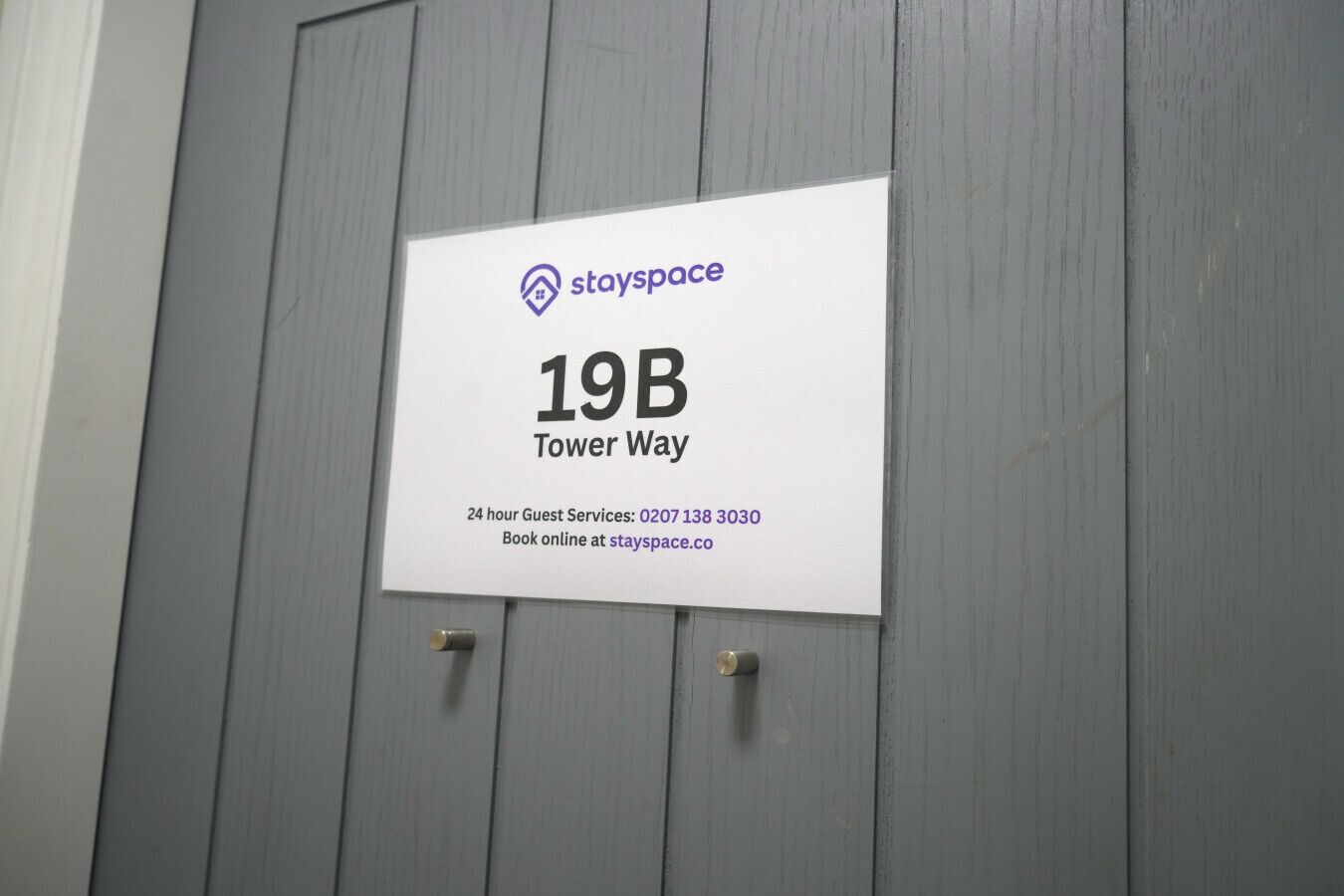 A sign on a grey door indicating '19B Tower Way' with the StaySpace logo, a 24-hour guest service number, and an online booking option.