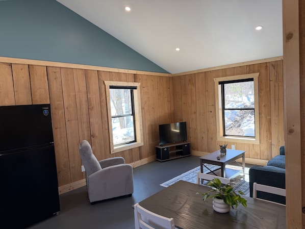 A room with wooden walls, a television on a stand, a dining table with chairs, and a refrigerator.