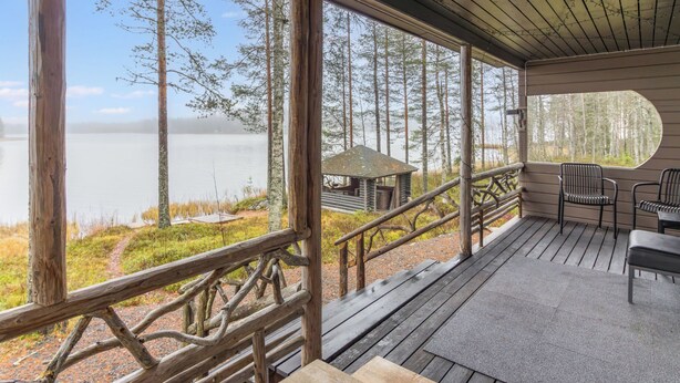 Una terraza de madera con vistas a un lago y un bosque, una cabaña pequeña y mobiliario para exteriores.