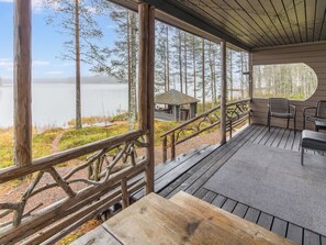 Una terraza de madera con vistas a un lago y un bosque, una cabaña pequeña y mobiliario para exteriores.