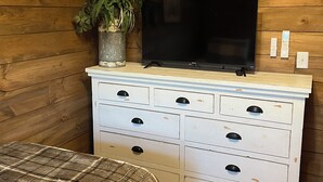 A wooden cabin room with a bed, a TV on a dresser, a plant, and a painting on the wall.