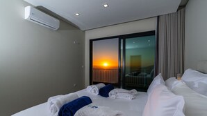 Une chambre d’hôtel avec un lit, deux serviettes enroulées, un appareil de climatisation, une table de chevet et une vue sur le coucher du soleil, visible à travers une porte coulissante.