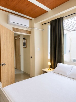 Ein Hotelzimmer mit einem Bett, einer Nachttischlampe, einer Klimaanlage, einem Fenster mit Vorhängen und einer Tür zu einem weiteren Raum.