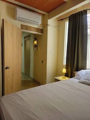 Une chambre d’hôtel comprenant un lit, une lampe de chevet, un appareil de climatisation, une fenêtre avec des rideaux et une porte donnant sur une autre pièce.