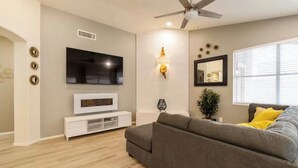 Ein modernes Wohnzimmer mit einem grauen Sofa, einem flachbildfernseher an der Wand, einem Deckenventilator und einem Fenster mit Jalousien.