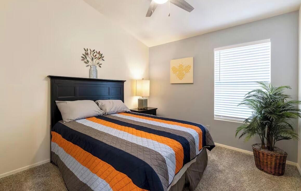 A bedroom with a bed, a nightstand, a lamp, a wall art piece, a window with blinds, and a potted plant.