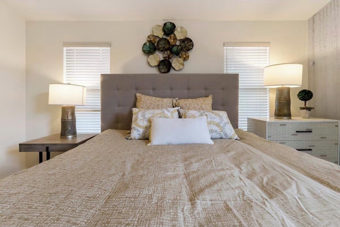 A bedroom with a large bed, two bedside tables with lamps, a decorative wall piece, and a window with blinds.