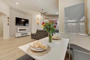 Un coin repas moderne avec une table blanche dressée pour deux personnes, un téléviseur à écran plat fixé au mur et un canapé gris en arrière-plan.