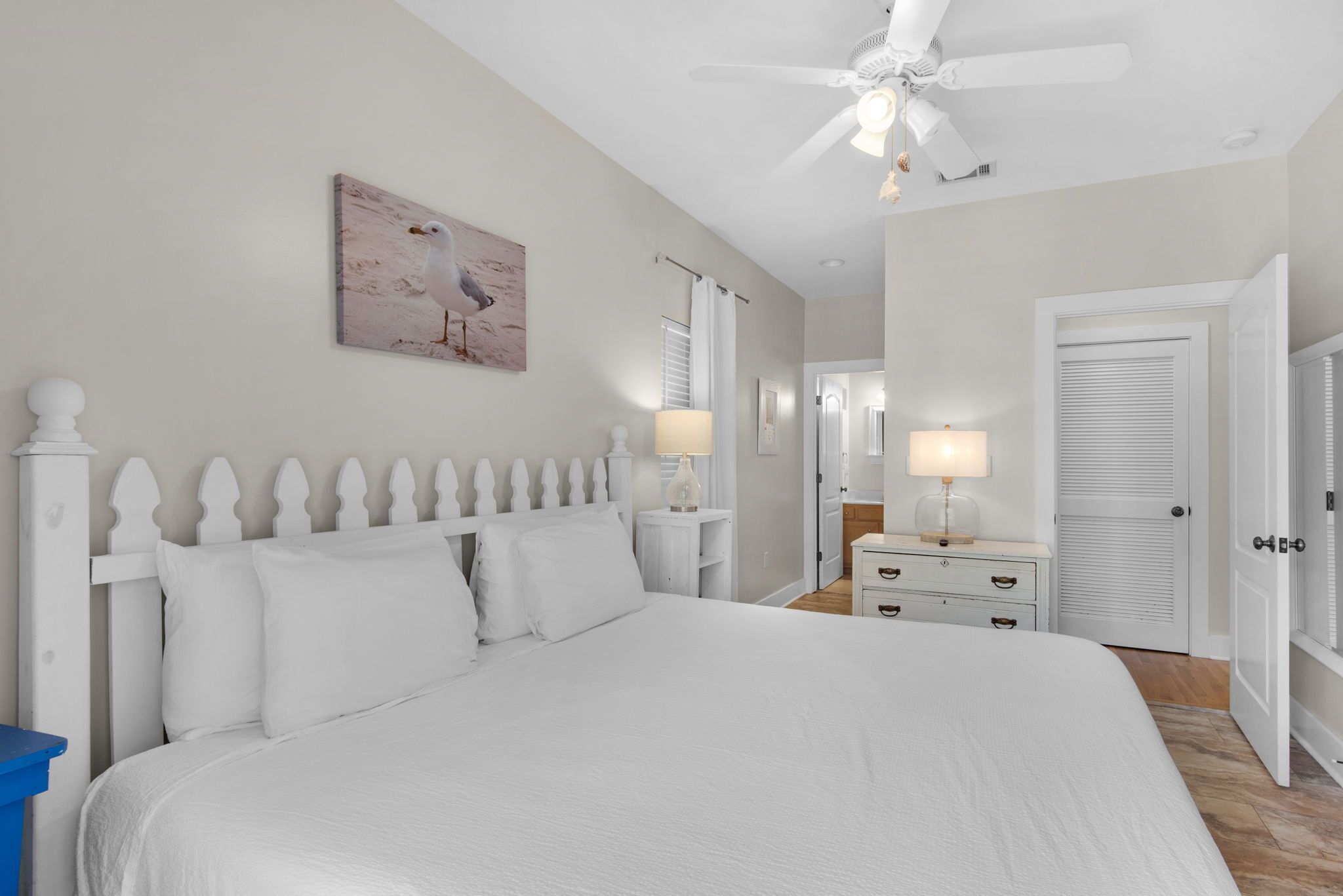 A bedroom with a white bed, a ceiling fan, a nightstand with a lamp, a dresser, and a door.