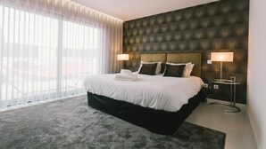 Een moderne hotelkamer met een groot bed, twee bedlampen, een donker hoofdeinde met structuur en uitzicht door dunne gordijnen.