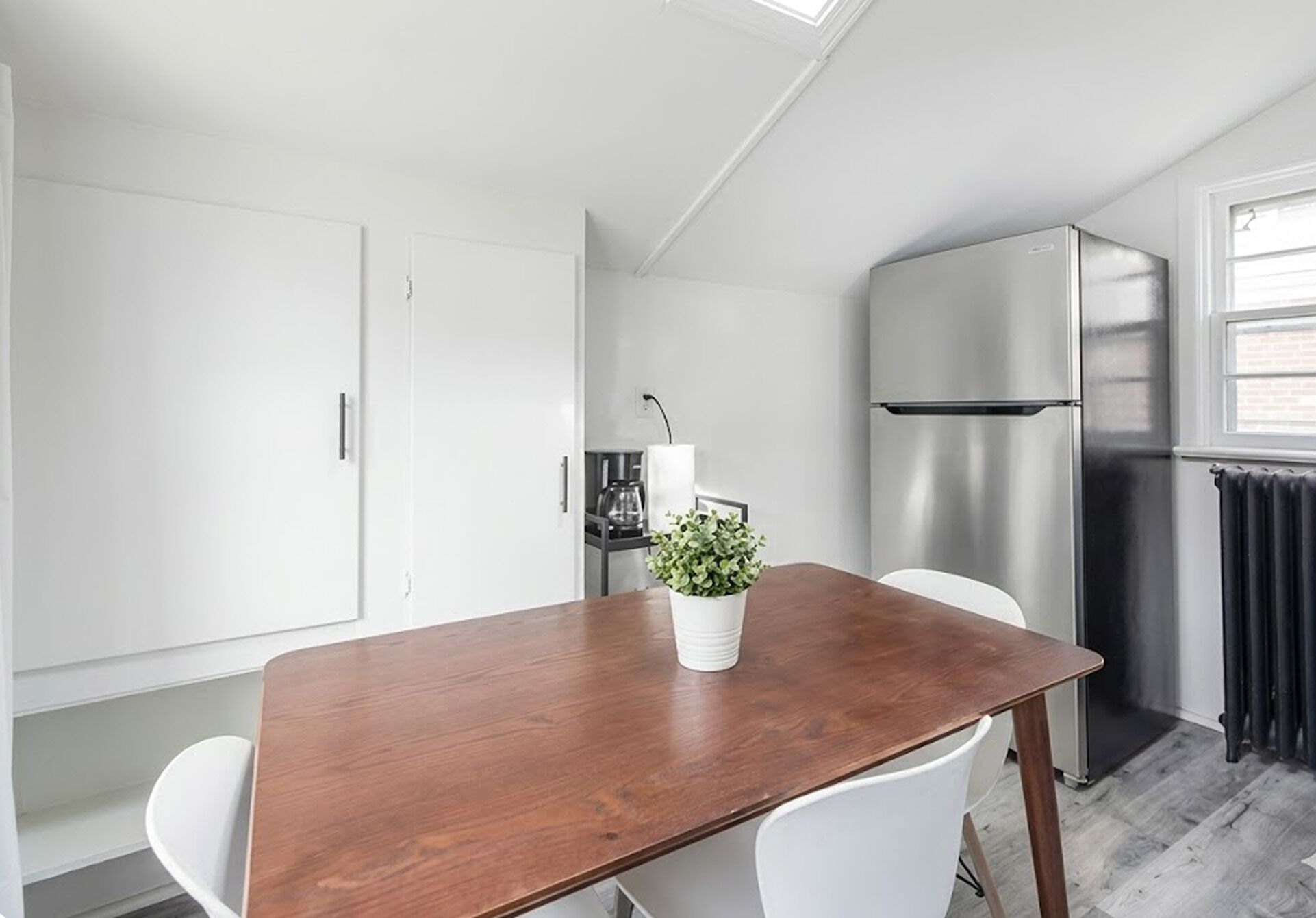 A compact kitchen with a wooden table, white chairs, a stainless steel refrigerator, and a potted plant.