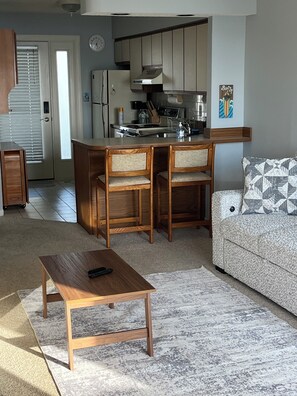 A living room with a wooden coffee table, two wooden chairs, a sofa with patterned cushions, a kitchen area with a stove and cabinets, and a clock on the wall.