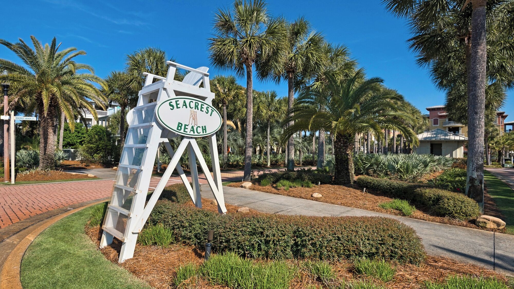 Naamplaatje voor Seacrest Beach met een witte achtergrond en groene letters, omgeven door palmbomen en een verharde looppad.