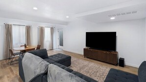 Ein modernes Wohnzimmer mit Flachbildfernseher, grauen Sofas, einem Holzschrank und einer Essecke mit Tisch und Stühlen.