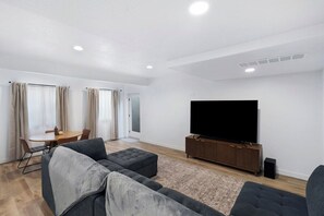 A modern living room with a flat-screen TV, grey sofas, a wooden cabinet, and a dining area with a table and chairs. - Lehi Basement: 2 Kings, 4 Queens, Pool & Parks (Lehi)